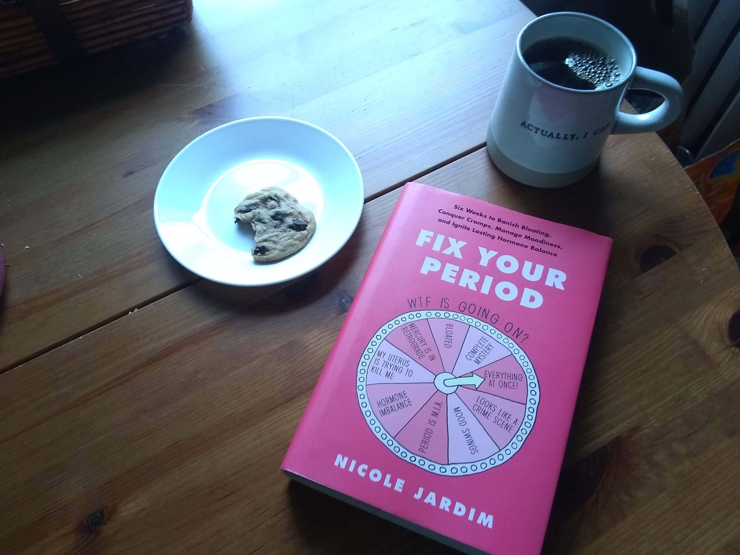 A copy of Fix your period on a wooden table with a white mug filled with coffee that says, "actually I can" and a partially eaten cookie